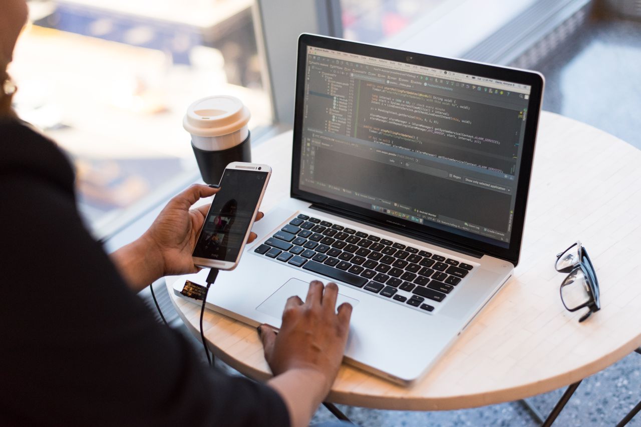 A developer holds an Android phone plugged into a laptop showing code in Android Studio, with a coffee cup and glasses on a round café table